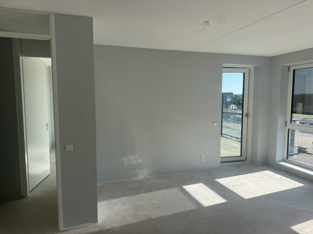 Kamer in een nieuwbouwappartement met lichtgrijze wanden, een balkondeur en een betonvloer